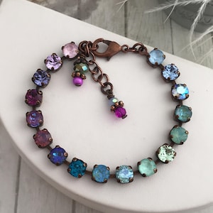 May include: A copper-toned bracelet featuring a series of colorful, square-cut crystals in shades of purple, blue, and green. The bracelet has a lobster clasp closure and a small chain extension. Additional small, dangling crystals add detail.