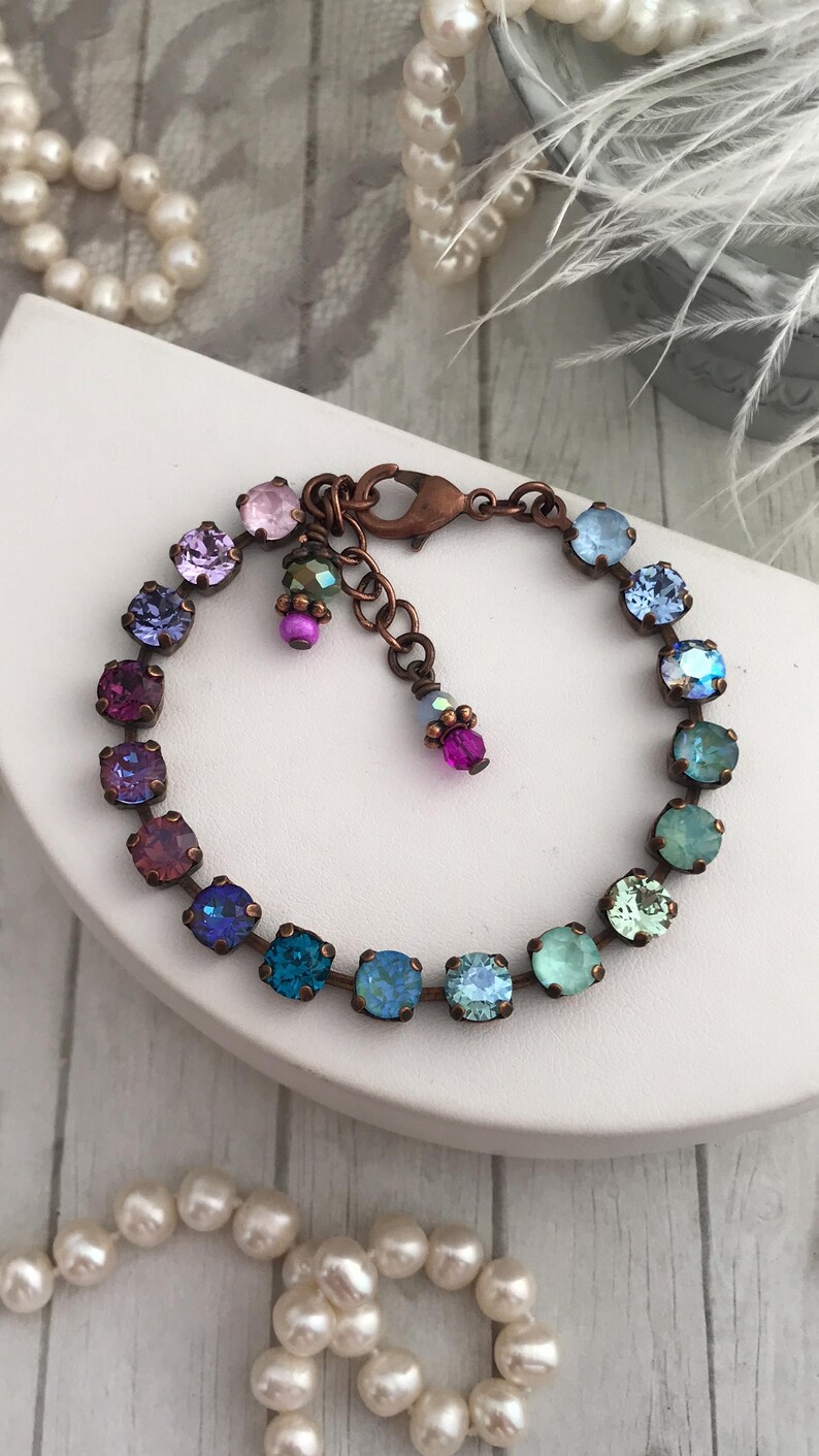 May include: A copper-toned bracelet featuring a series of colorful, square-cut crystals in shades of purple, blue, and green. The bracelet has a lobster clasp closure and a small chain extension. Additional small, dangling crystals add detail.