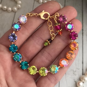 May include: A gold-toned bracelet featuring a series of colorful, iridescent crystals. The crystals transition through shades of purple, teal, yellow, orange, and pink. The bracelet has a lobster clasp closure and a small chain for adjustable sizing.