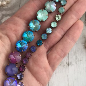 May include: A collection of colorful, iridescent crystal jewelry pieces in shades of blue, green, purple, and pink. The crystals are set in bronze-colored metal. A strand of pearls is visible in the background.