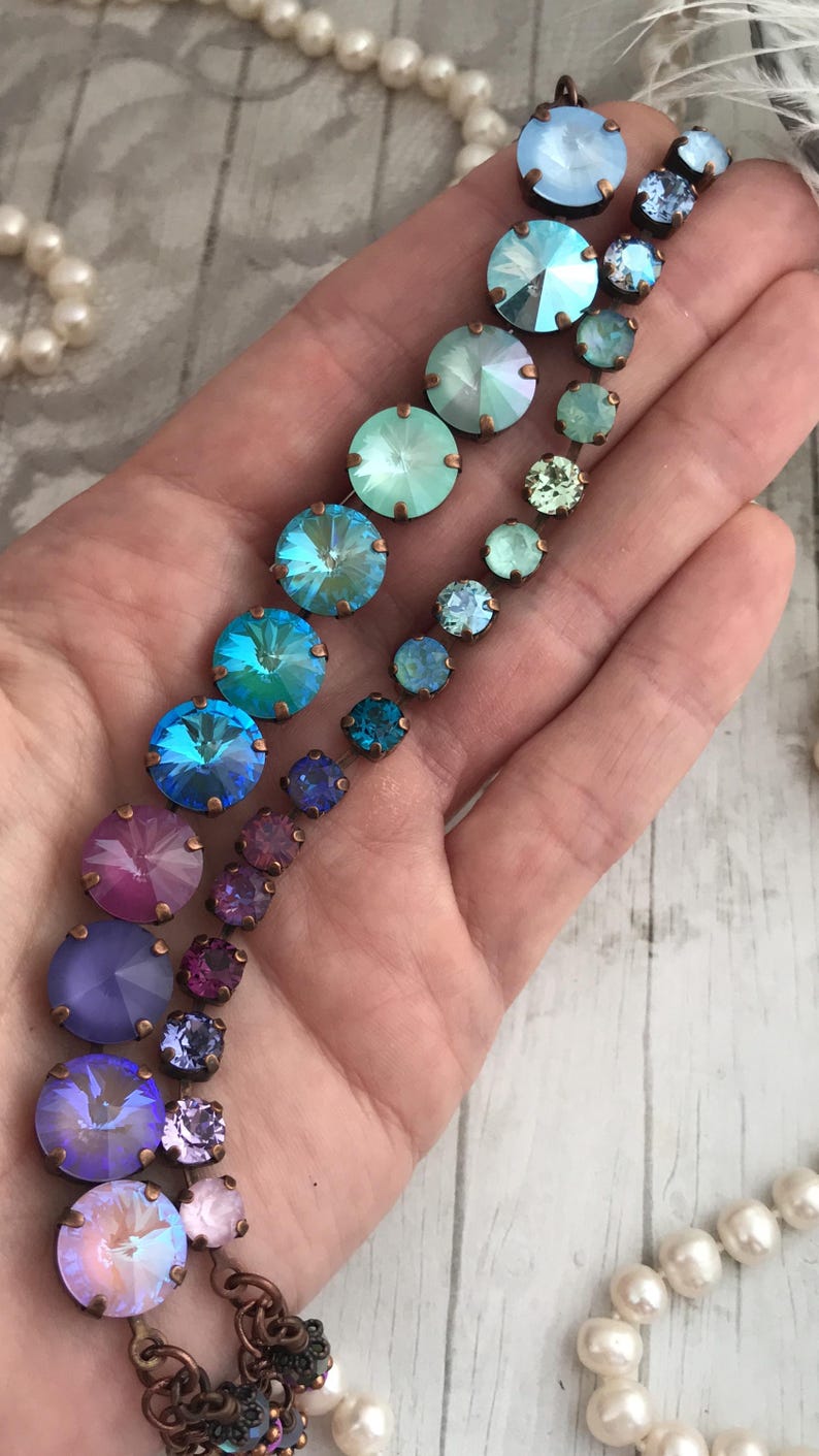 May include: A collection of colorful, iridescent crystal jewelry pieces in shades of blue, green, purple, and pink. The crystals are set in bronze-colored metal. A strand of pearls is visible in the background.