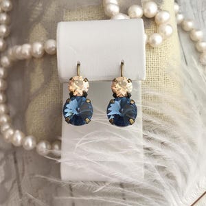 May include: A pair of dangle earrings with two crystal stones. The top stone is a light peach colour and the bottom stone is a dark blue colour. The earrings are on a white display card.