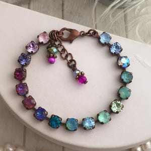 May include: A copper-toned bracelet featuring a series of colorful, square-cut gemstones in shades of pink, purple, blue, and green. The bracelet has a delicate chain and a lobster clasp closure. Additional small, dangling beads add detail.