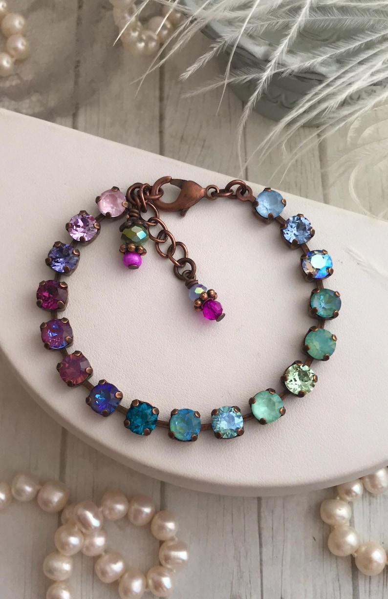 May include: A copper-toned bracelet featuring a series of colorful, square-cut gemstones in shades of pink, purple, blue, and green. The bracelet has a delicate chain and a lobster clasp closure. Additional small, dangling beads add detail.