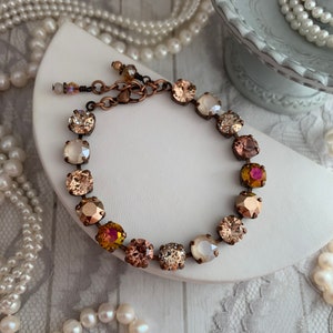 May include: A copper chain bracelet with a variety of crystal beads in shades of brown, pink, and white. The bracelet is displayed on a white surface with a pearl necklace in the background.
