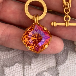 Pode incluir: Um colar de corrente de ouro com um grande pingente de pedra preciosa facetada laranja e roxo. O pingente est&aacute; preso a um anel de ouro com uma borda decorativa.