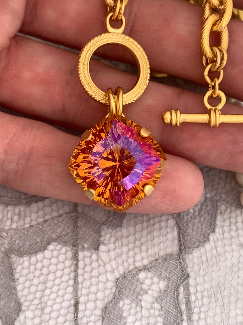 Pode incluir: Um colar de corrente de ouro com um grande pingente de pedra preciosa facetada laranja e roxo. O pingente est&aacute; preso a um anel de ouro com uma borda decorativa.