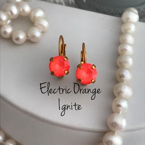 May include: A pair of gold hoop earrings with bright orange round crystal stones. The earrings are on a white surface with a pearl necklace. The text "Electric Orange Ignite" is below the earrings.