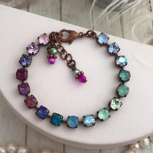 May include: A multi-colored bracelet featuring square-cut crystals in shades of pink, purple, blue, and green. The crystals are set in a copper-toned metal, with a matching clasp and decorative dangles. The bracelet is displayed on a white surface.