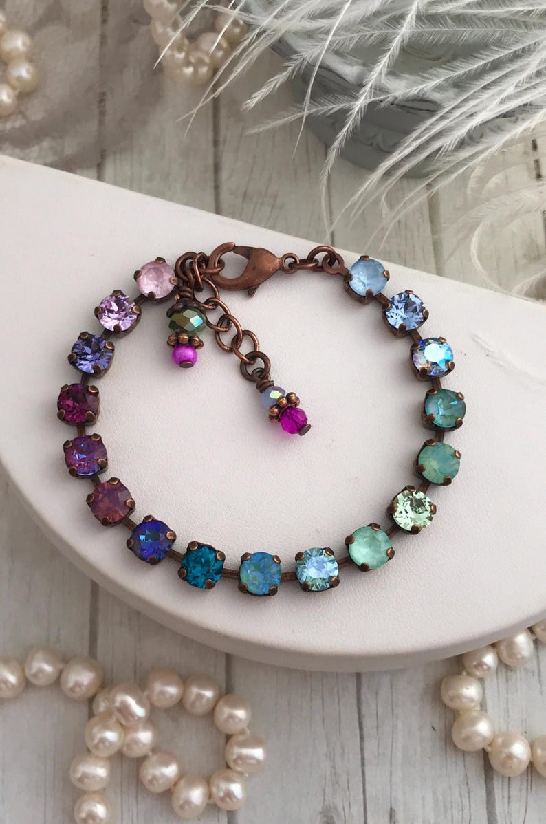 May include: A multi-colored bracelet featuring square-cut crystals in shades of pink, purple, blue, and green. The crystals are set in a copper-toned metal, with a matching clasp and decorative dangles. The bracelet is displayed on a white surface.