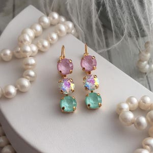 May include: A pair of gold dangle earrings with three crystals each. The top crystal is pink, the middle crystal is iridescent, and the bottom crystal is light green.