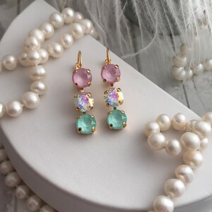 May include: A pair of gold dangle earrings with three round crystals. The top crystal is pink, the middle crystal is iridescent, and the bottom crystal is light green.