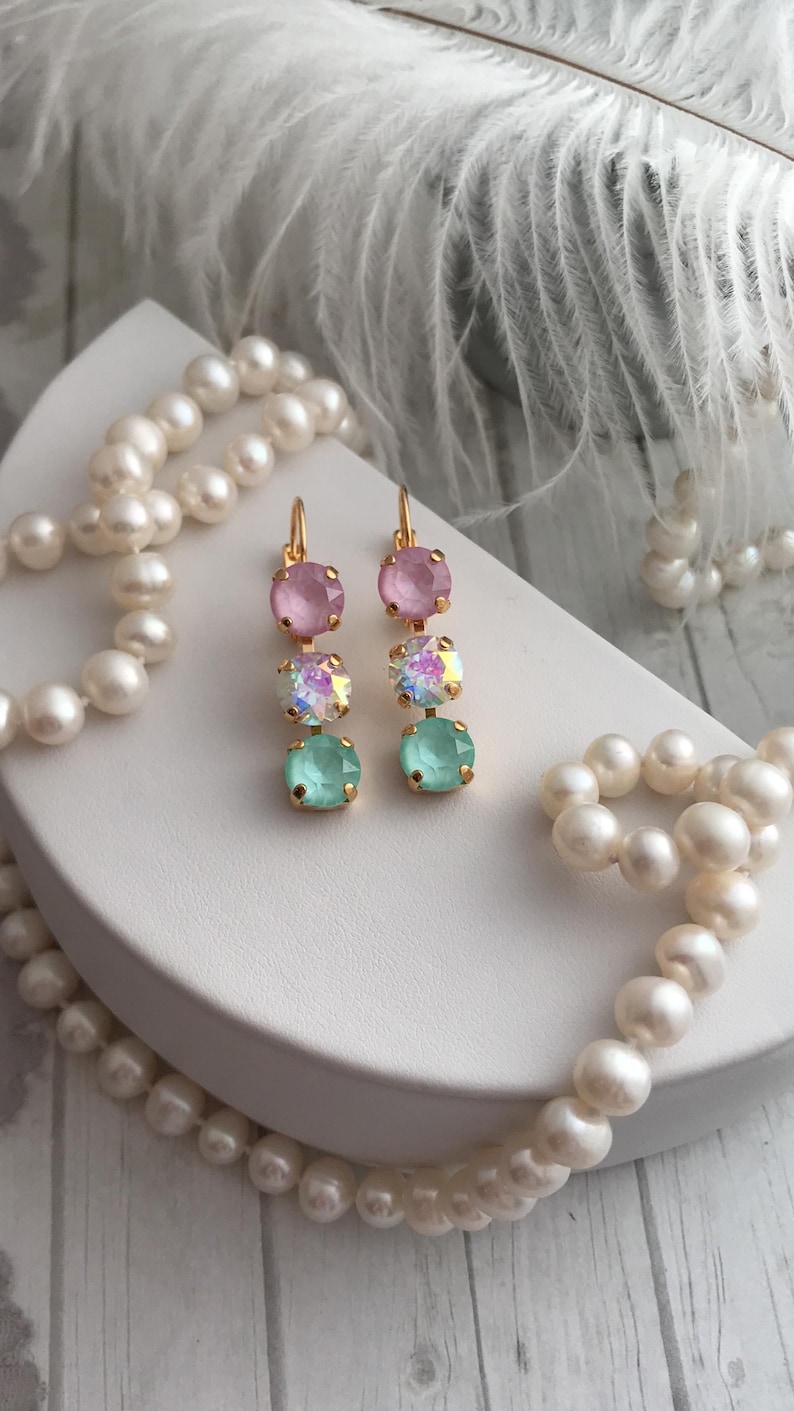 May include: A pair of gold dangle earrings with three round crystals. The top crystal is pink, the middle crystal is iridescent, and the bottom crystal is light green.