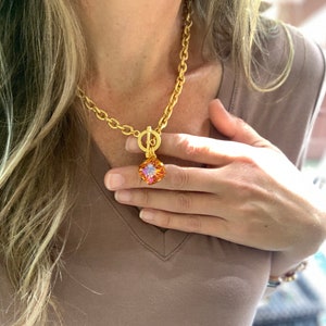 Pode incluir: Um colar de corrente de ouro com fecho de trava e um pingente quadrado com uma gema multicolorida.