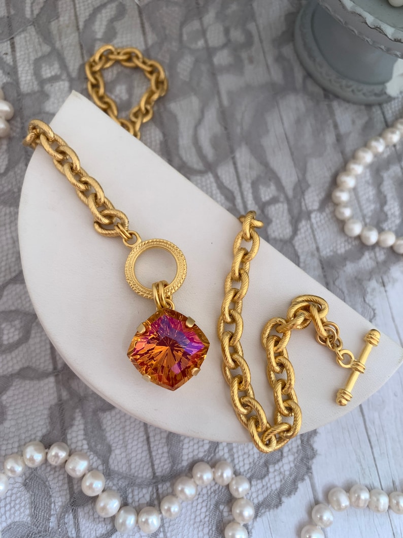 Pode incluir: Um colar de corrente de ouro com um grande pingente de pedra preciosa facetada laranja e rosa. A corrente tem um design texturizado tipo corda e um simples anel redondo de ouro na parte superior do pingente.