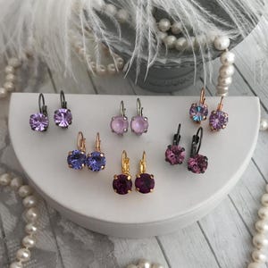 May include: Six pairs of earrings with different colored crystal stones. The earrings are silver, gold, and black with purple, pink, and lavender stones.