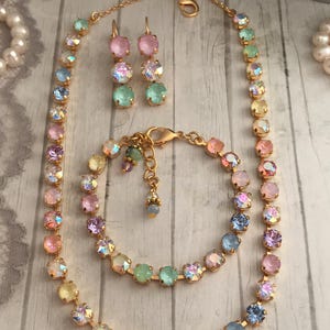 May include: A set of pastel rainbow colored crystal jewelry, including a necklace, bracelet, and earrings. The jewelry is made with gold-toned metal and features iridescent crystals.