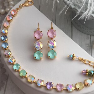 May include: A gold bracelet and earrings set with pastel colored crystals. The bracelet has a rainbow gradient of crystals, including pink, blue, green, purple, and yellow. The earrings have three crystals each, in pink, iridescent, and green.