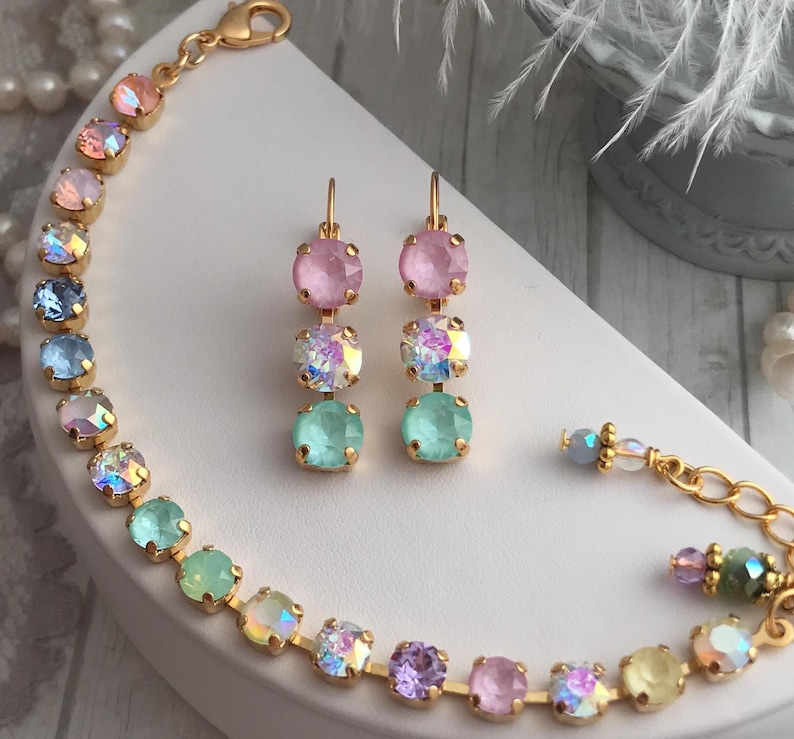 May include: A gold bracelet and earrings set with pastel colored crystals. The bracelet has a rainbow gradient of crystals, including pink, blue, green, purple, and yellow. The earrings have three crystals each, in pink, iridescent, and green.