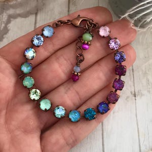 May include: A colorful bracelet featuring a series of iridescent, round gemstones in shades of blue, green, purple, and pink. The stones are set in a copper-colored metal and connected by a delicate chain. The bracelet is approximately 7 inches in length.