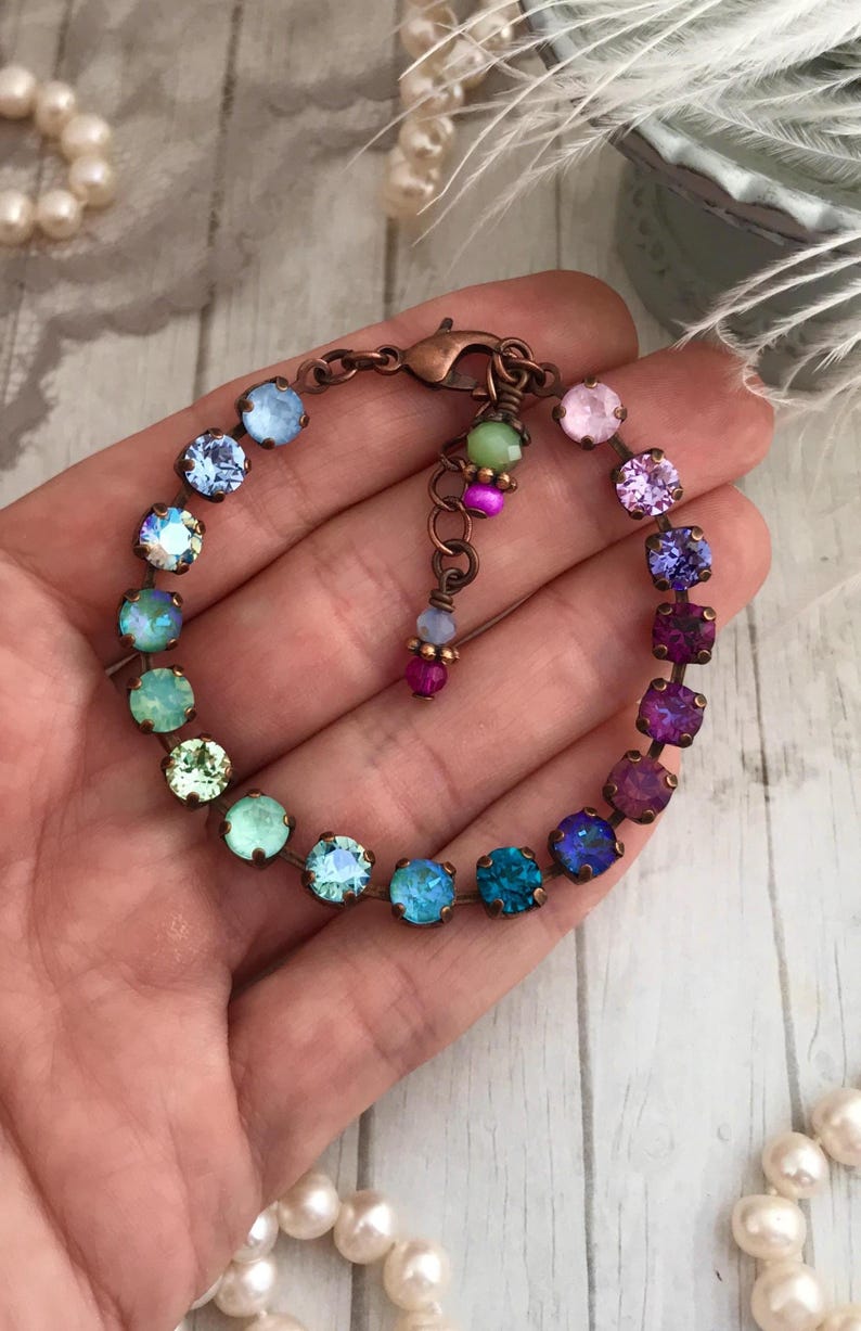 May include: A colorful bracelet featuring a series of iridescent, round gemstones in shades of blue, green, purple, and pink. The stones are set in a copper-colored metal and connected by a delicate chain. The bracelet is approximately 7 inches in length.