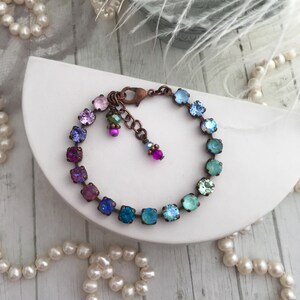 May include: A copper-toned bracelet featuring a gradient of colorful square crystals. The bracelet transitions from pink to blue-green hues. Additional small, dangling charms with pink and green beads add detail. The bracelet is displayed on a white surface with pearl strands.
