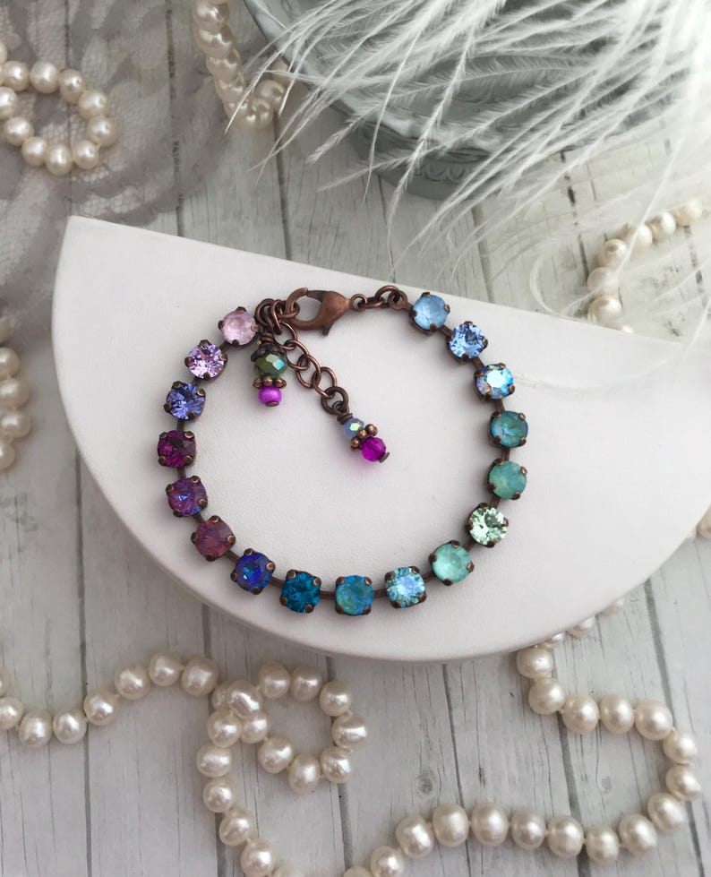 May include: A copper-toned bracelet featuring a gradient of colorful square crystals. The bracelet transitions from pink to blue-green hues. Additional small, dangling charms with pink and green beads add detail. The bracelet is displayed on a white surface with pearl strands.