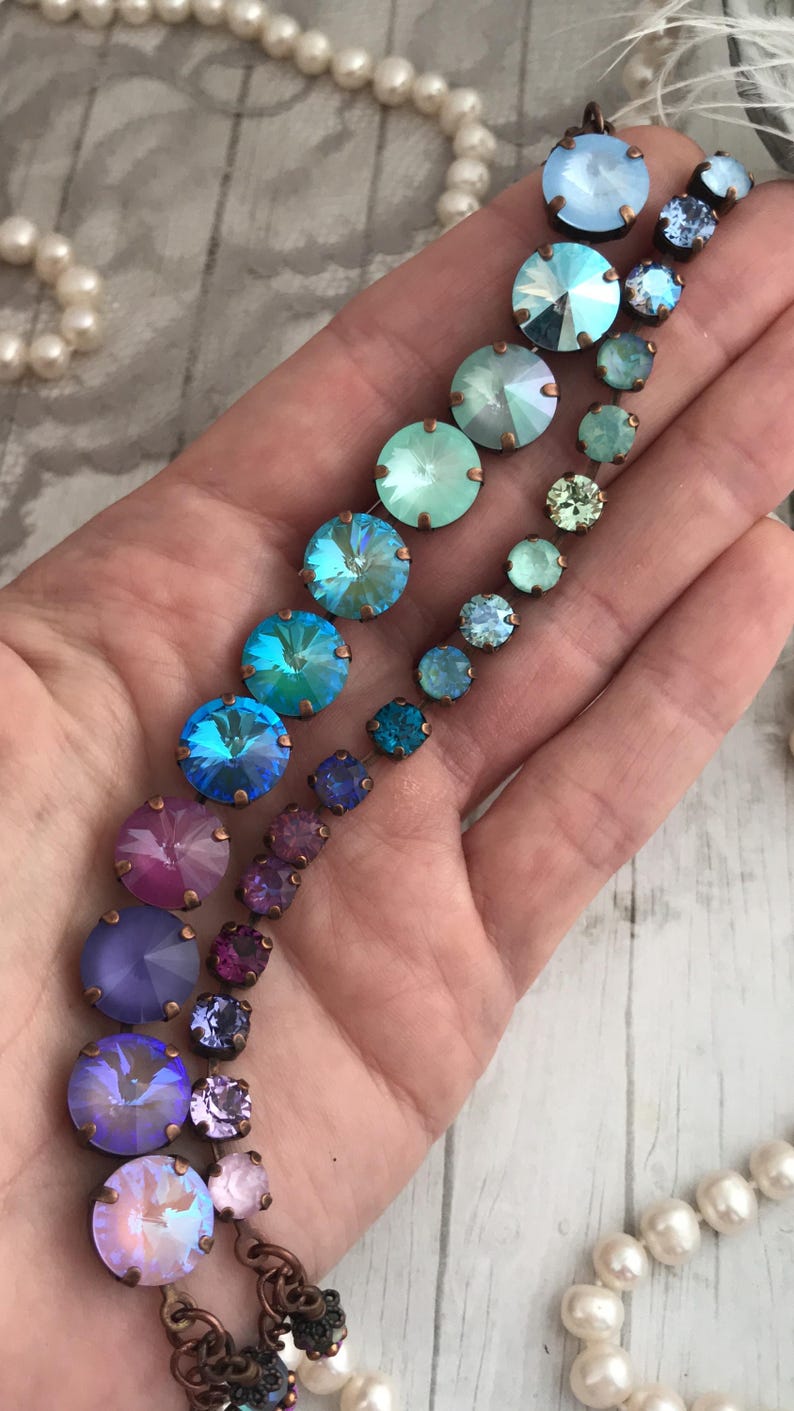 May include: A collection of iridescent crystal jewelry in shades of blue, purple, and pink. The crystals are set in copper-colored metal and arranged in a cascading design. A pearl necklace is visible in the background.