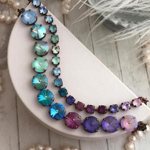 May include: Two rows of colorful crystal jewelry are displayed on a white, crescent-shaped stand. The top row features small, square crystals in shades of blue and green. The bottom row has larger, round crystals in shades of blue, purple, and pink.