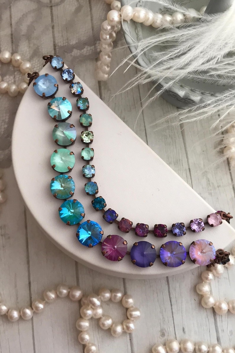 May include: Two rows of colorful crystal jewelry are displayed on a white, crescent-shaped stand. The top row features small, square crystals in shades of blue and green. The bottom row has larger, round crystals in shades of blue, purple, and pink.