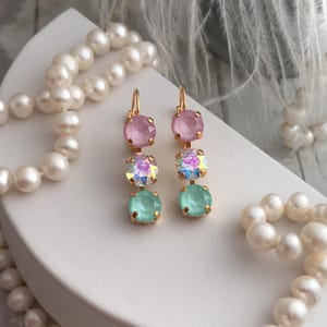 May include: A pair of gold dangle earrings with three crystal stones each. The top stone is pink, the middle stone is iridescent, and the bottom stone is light green.