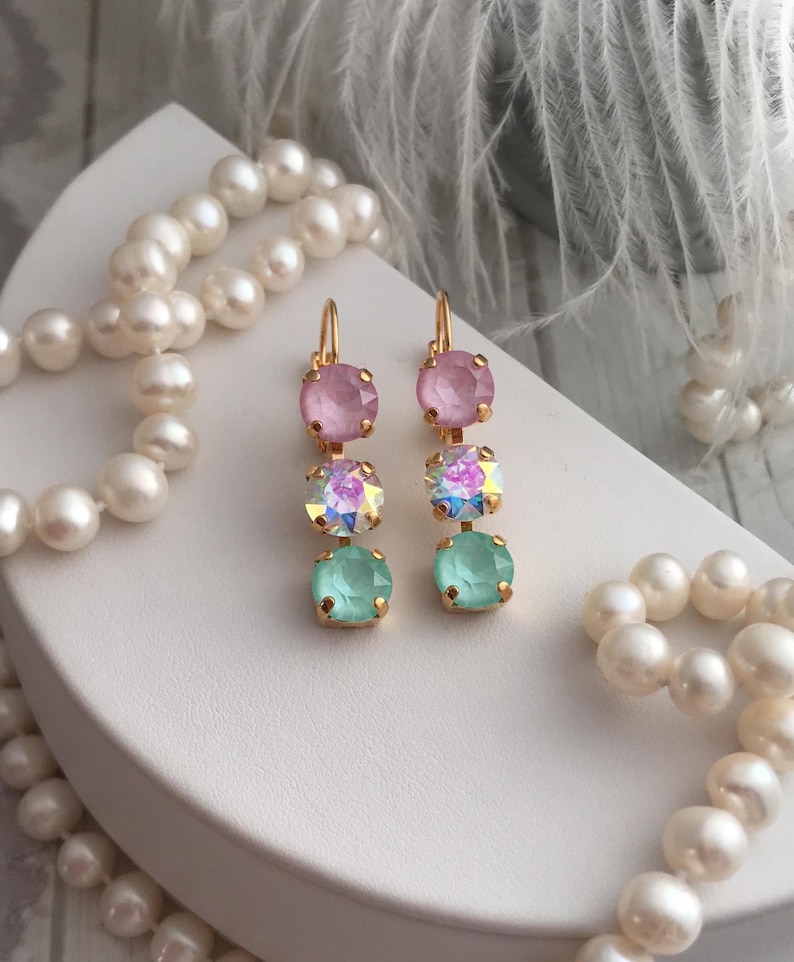 May include: A pair of gold dangle earrings with three crystal stones each. The top stone is pink, the middle stone is iridescent, and the bottom stone is light green.