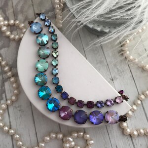 May include: Two necklaces with iridescent, round and square crystals in shades of blue, teal, purple, and pink. The crystals are set in a bronze-colored metal. The necklaces are displayed on a white, crescent-shaped surface, with pearls and feathers in the background.
