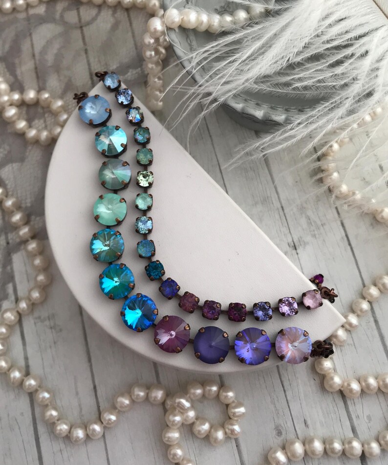 May include: Two necklaces with iridescent, round and square crystals in shades of blue, teal, purple, and pink. The crystals are set in a bronze-colored metal. The necklaces are displayed on a white, crescent-shaped surface, with pearls and feathers in the background.