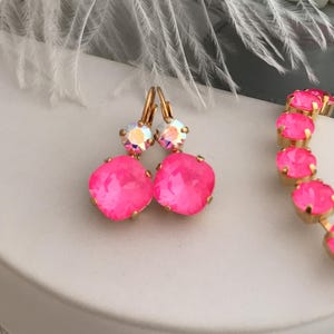 Könnte beinhalten: Ein Paar rosa Kristallohrringe mit goldfarbenen Haken und einem kleineren, klaren Kristall-Akzent. Die Ohrringe werden auf einer weißen Oberfläche präsentiert.