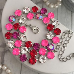 May include: A sparkling bracelet featuring a series of round, colorful rhinestones in shades of pink, red, and clear. The bracelet has a silver-tone chain and clasp, and is displayed on a white surface. Pearls are in the background.