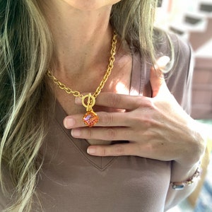 Pode incluir: Um colar de corrente de ouro com um grande pingente quadrado laranja. O pingente &eacute; feito de vidro facetado e tem um fecho dourado.