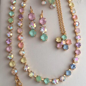 May include: A set of pastel-colored crystal jewelry featuring a necklace, earrings, and a pendant. The necklace is a double strand of gold chain with pastel-colored crystals. The earrings are a pair of drop earrings with pastel-colored crystals. The pendant is a flower-shaped charm with pastel-colored crystals.