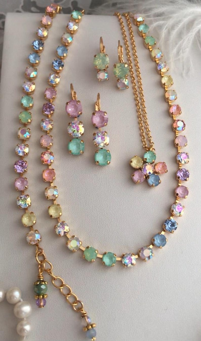 May include: A set of pastel-colored crystal jewelry featuring a necklace, earrings, and a pendant. The necklace is a double strand of gold chain with pastel-colored crystals. The earrings are a pair of drop earrings with pastel-colored crystals. The pendant is a flower-shaped charm with pastel-colored crystals.