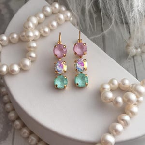 May include: A pair of gold dangle earrings with three crystal stones each. The top stone is pink, the middle stone is iridescent, and the bottom stone is light green.