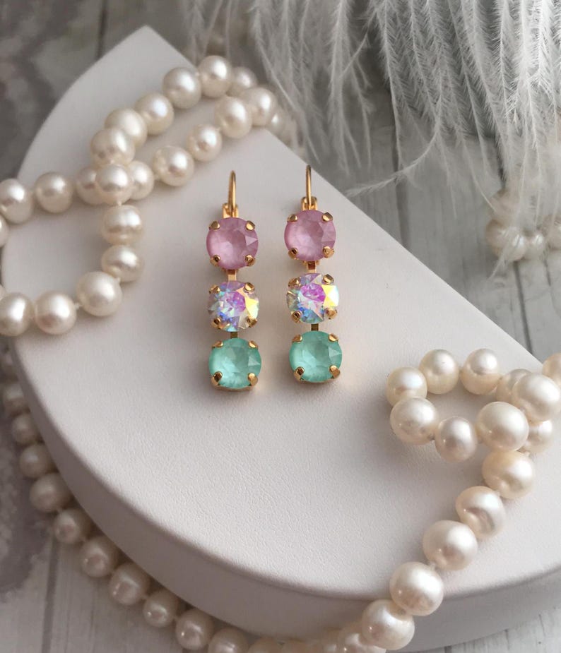 May include: A pair of gold dangle earrings with three crystal stones each. The top stone is pink, the middle stone is iridescent, and the bottom stone is light green.