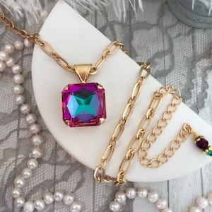 May include: A gold-toned necklace with a large, square, iridescent pendant in shades of pink, purple, and teal. The necklace features a chunky chain and is displayed with a pearl necklace and other jewelry.