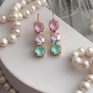 May include: A pair of gold earrings with three crystal stones each. The top stone is pink, the middle stone is iridescent, and the bottom stone is light blue.