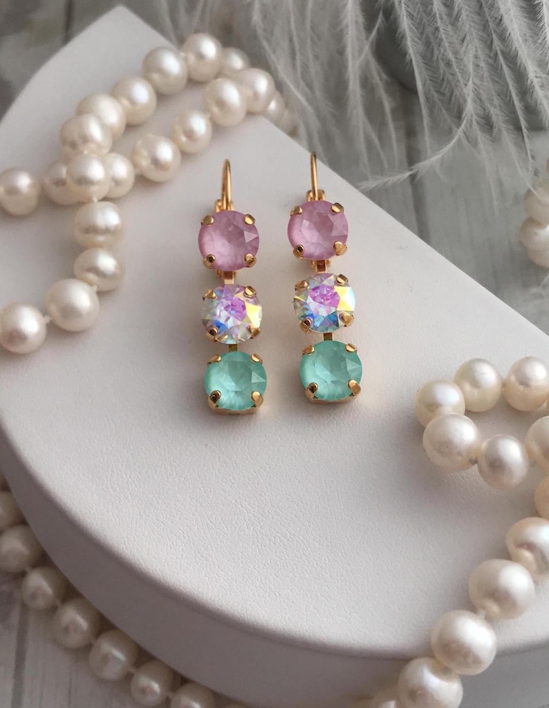 May include: A pair of gold earrings with three crystal stones each. The top stone is pink, the middle stone is iridescent, and the bottom stone is light blue.