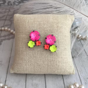 May include: A pair of vibrant, neon-colored earrings. Each earring features a large, hot pink crystal surrounded by smaller orange and chartreuse crystals. The earrings are displayed on a small, beige burlap pillow.