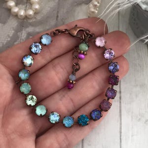 May include: A multi-colored crystal bracelet with a copper-colored chain. The bracelet features a variety of round crystals in shades of blue, green, purple, and pink. The bracelet is held in a hand.