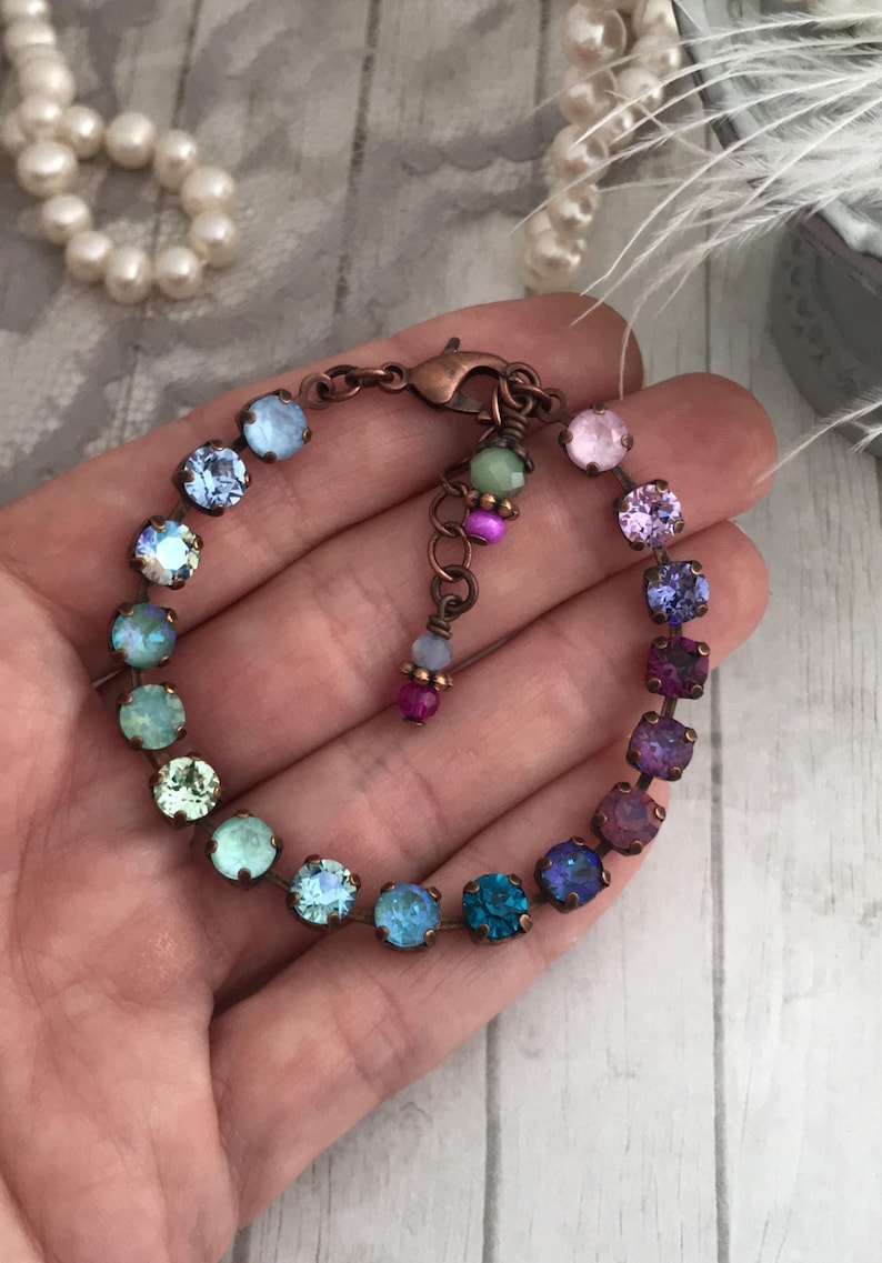 May include: A multi-colored crystal bracelet with a copper-colored chain. The bracelet features a variety of round crystals in shades of blue, green, purple, and pink. The bracelet is held in a hand.