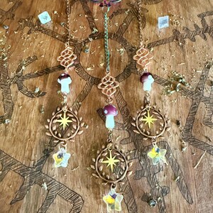 May include: Decorative hanging ornaments with copper-colored chains, featuring mushroom and star charms. The ornaments hang from a wooden surface with etched tree designs. The charms include clear and purple crystals.