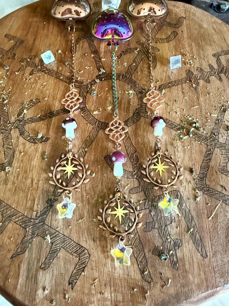 May include: Decorative hanging ornaments with copper-colored chains, featuring mushroom and star charms. The ornaments hang from a wooden surface with etched tree designs. The charms include clear and purple crystals.