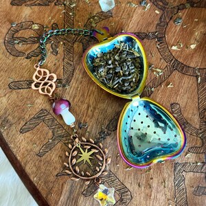 May include: Iridescent heart-shaped tea infuser filled with loose tea. The infuser is open, revealing the tea leaves. A chain with decorative charms, including a mushroom, star, and a crystal star, hangs from the infuser. The background is a wooden surface.